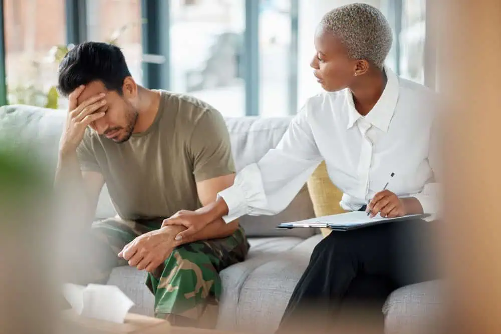 A woman in a white shirt sits on a couch, holding a clipboard and comforting a man in a green t-shirt and camouflage pants who appears distressed, with his head in his hand. They are in a well-lit room with large windows, typical of family counseling sessions in Fort Bend County.