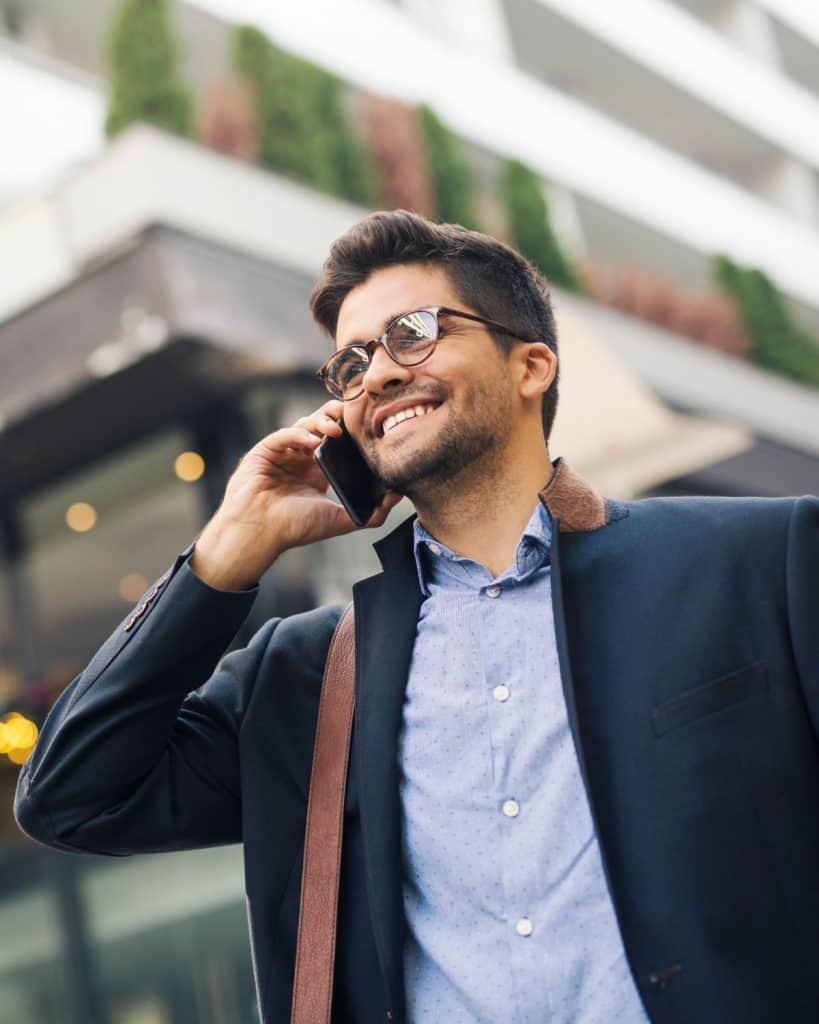 A man wearing glasses and a blazer smiles while talking on his smartphone. He stands outdoors in an urban setting with buildings and greenery, exuding the calm confidence of a therapist from Fort Bend County, TX.