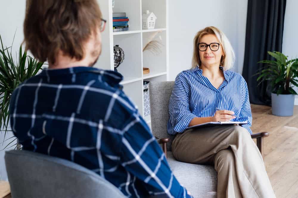A woman in a blue striped shirt and glasses is sitting in a chair, holding a clipboard and pen, attentively listening to a person in a plaid shirt. This serene setting, complete with bookshelves and plants, reflects the therapeutic ambiance typical of Fort Bend County, TX.