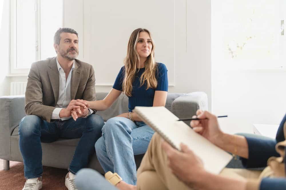 A man and woman sit on a couch holding hands, looking attentively at an unseen person during a counseling session in Fort Bend County, TX. The blurred figure in the foreground holds a notepad and pen, capturing the supportive atmosphere of therapy in the brightly lit room.