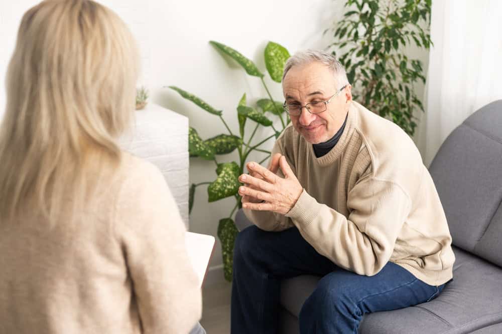 An elderly man in a beige sweater sits on a gray sofa, leaning forward with hands clasped, deep in conversation about therapy Fort Bend County TX. He talks to a woman with long blonde hair seated across from him, while a green plant adds warmth to the setting.