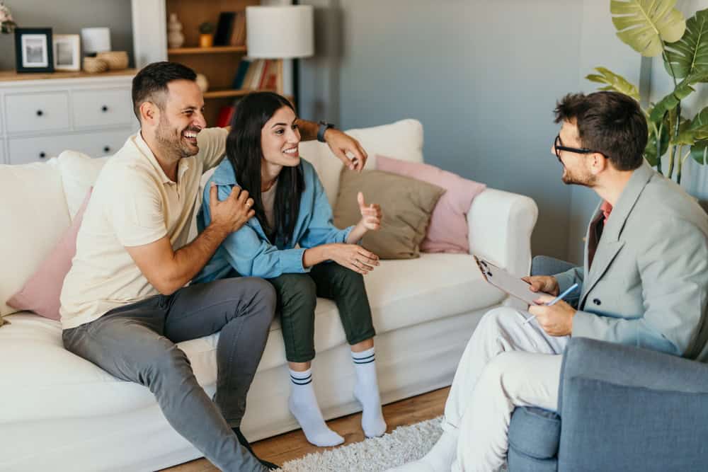 A couple sits on a sofa, smiling and engaged in conversation with a psychotherapist Fort Bend County TX, who is seated in an armchair. The setting appears to be a warmly decorated living room or office space.