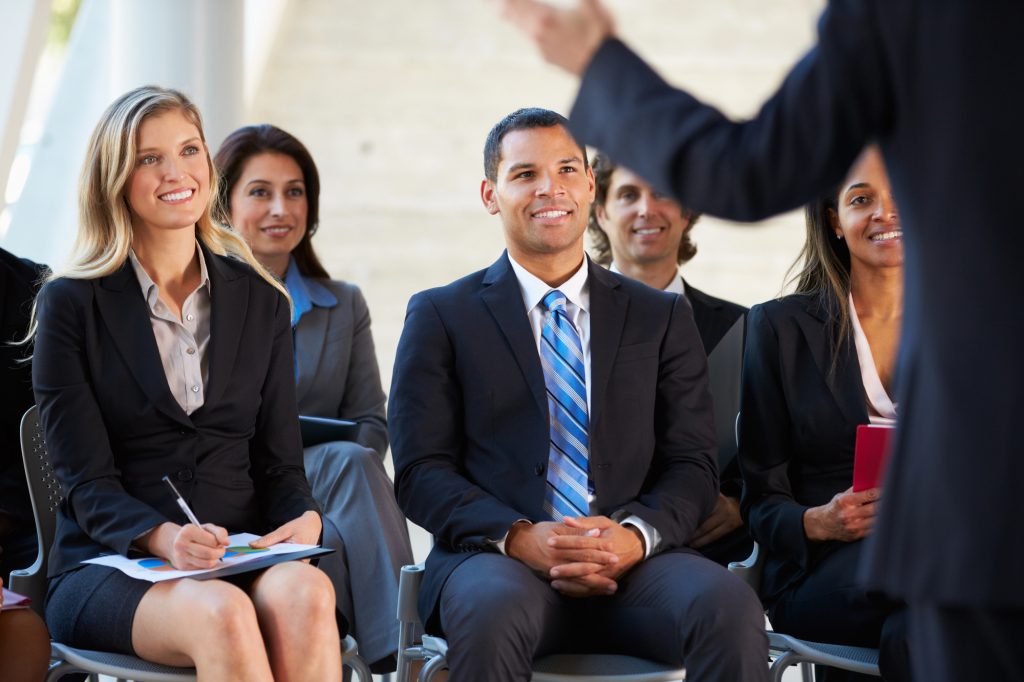 A group of professionally dressed adults sit and listen attentively to a speaker at a business seminar. Some are smiling, and one person is taking notes with a notepad and pen.