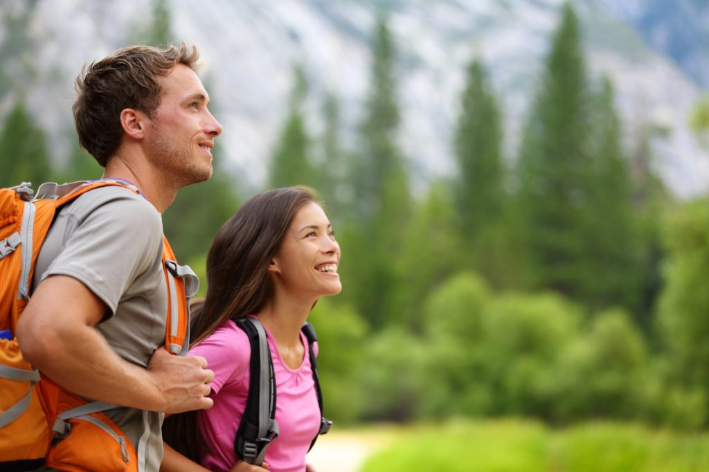 Two hikers, a man and a woman, wearing backpacks, stand outdoors smiling and looking upwards, with a blurred background of green forest and mountains.
